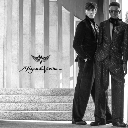 Two men in elegant tuxedos pose in a stylish architectural setting, highlighting sophisticated fashion against a textured backdrop.