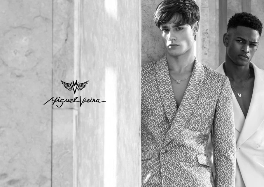 Two male models in a black and white portrait. One man wears a patterned blazer and a necklace, and the other wears a white suit.