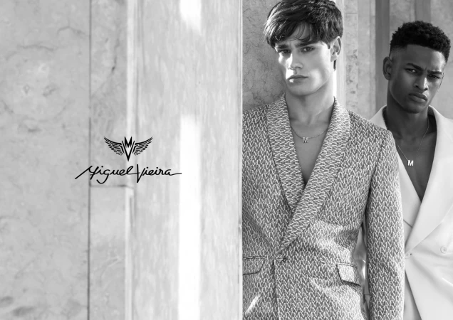 Two male models in a black and white portrait. One man wears a patterned blazer and a necklace, and the other wears a white suit.