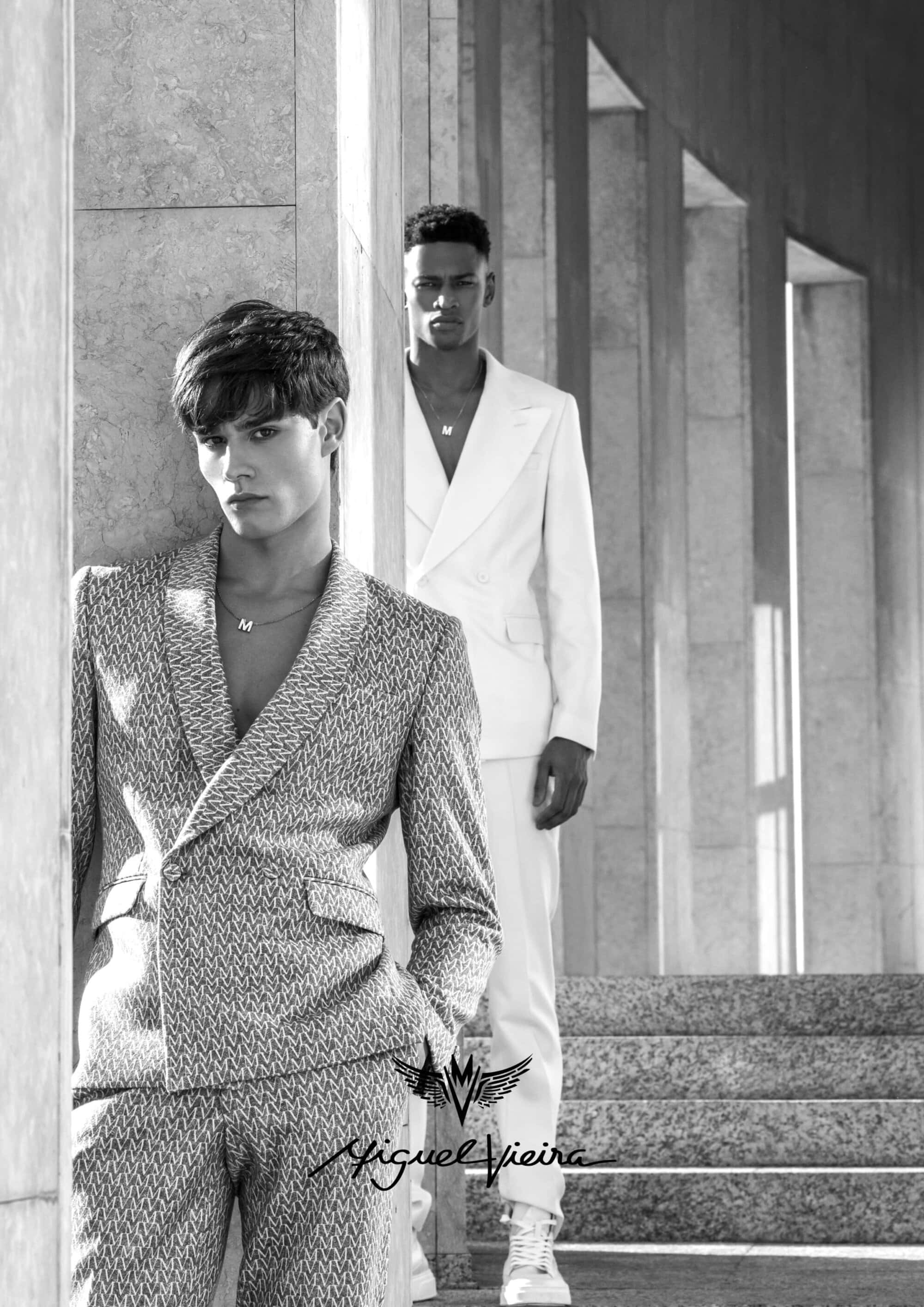 Two male fashion models pose on marble steps wearing patterned and solid suits near fluted columns in black and white monochrome.