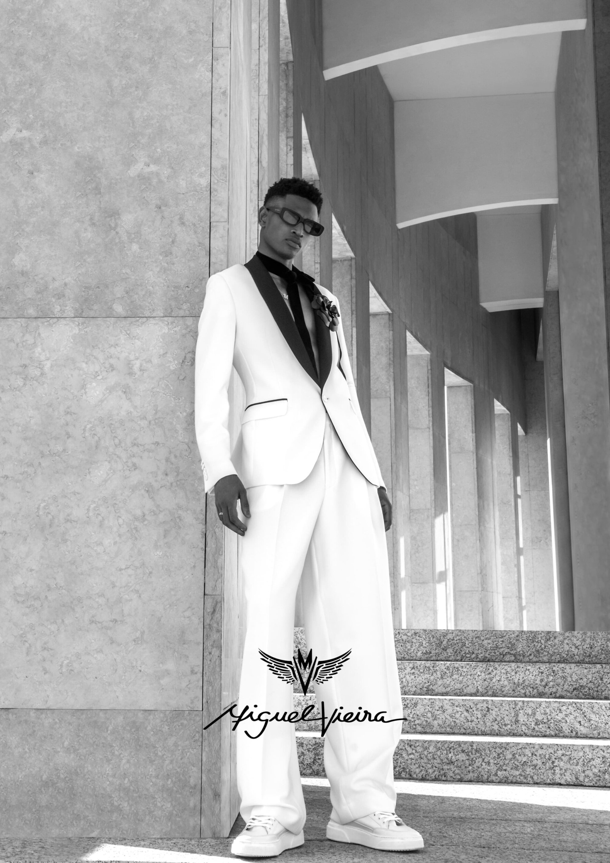Black and white portrait of an adult male posing in a white suit with a black lapel, sunglasses, and white sneakers outdoors.