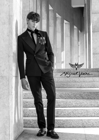 Black and white portrait of a male fashion model in a black suit, leaning against a wall with stairs in the background, showcasing formal wear.