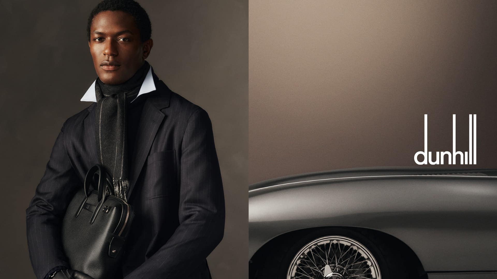 An African American man in a dark suit and scarf holds a black leather bag, beside a classic car, against a two-tone brown backdrop.