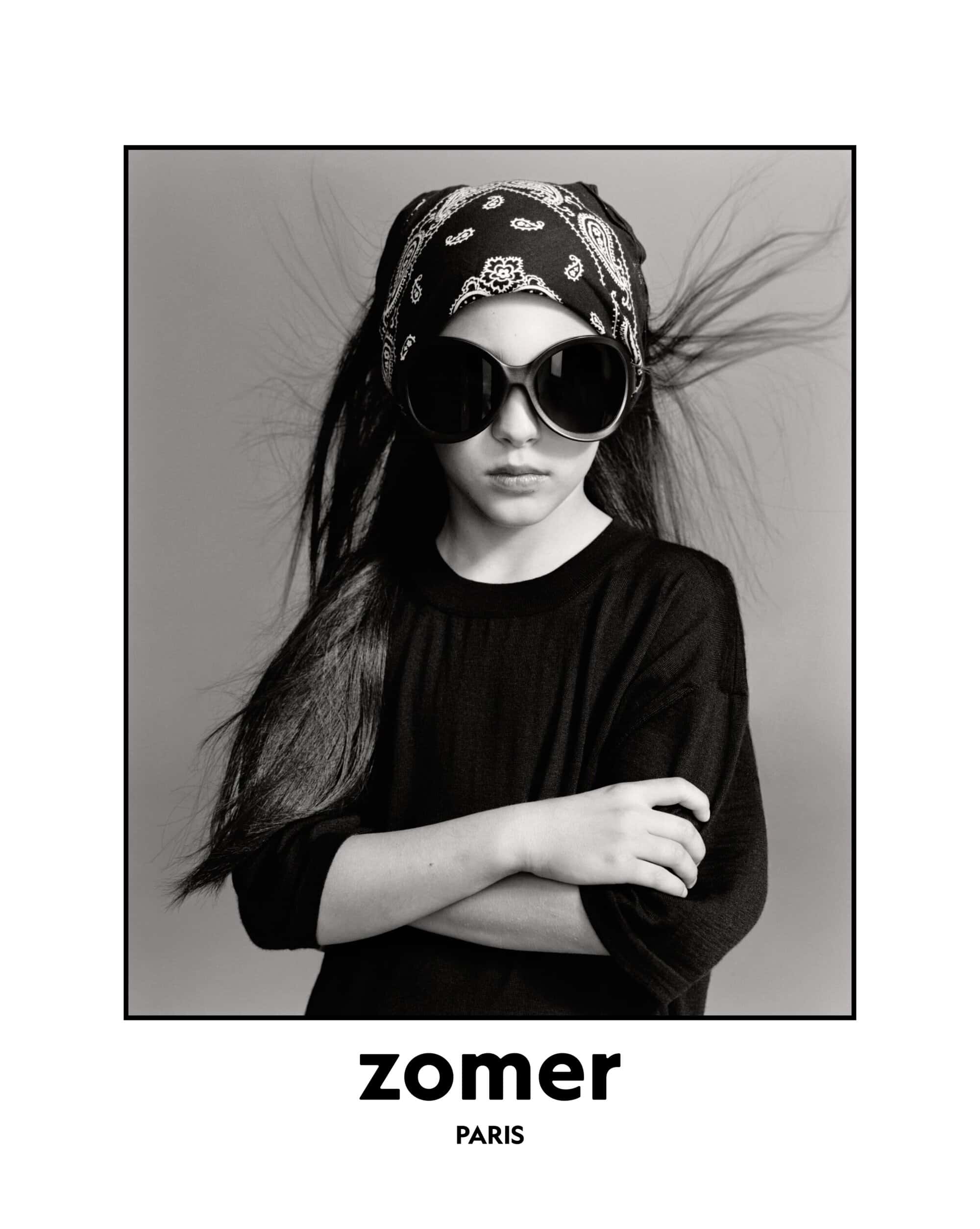 Black and white portrait of a child wearing oversized dark sunglasses and a patterned bandana headscarf with arms crossed.