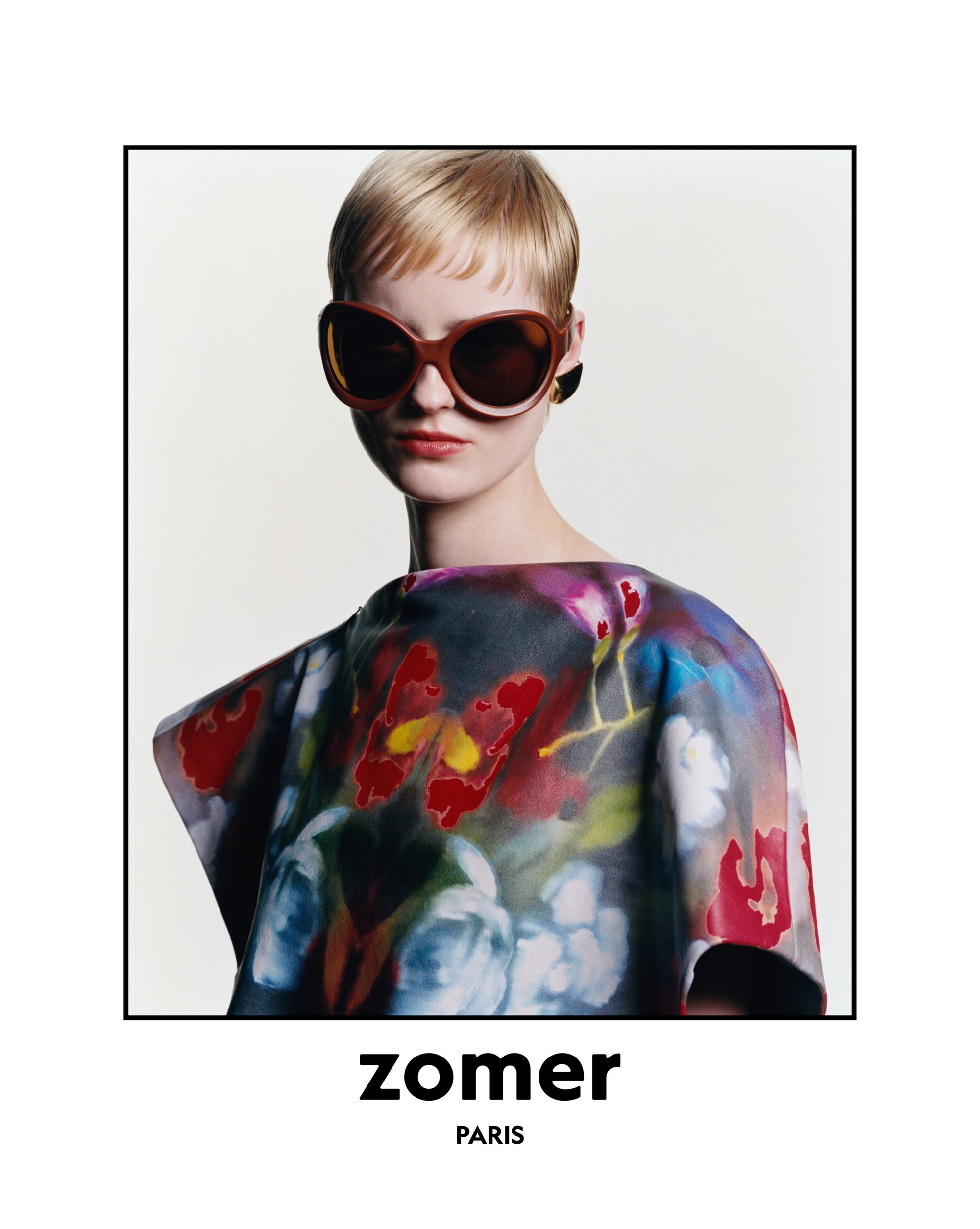 Fashion shot of a woman with short hair wearing sunglasses and a floral print top, posing against a white backdrop.