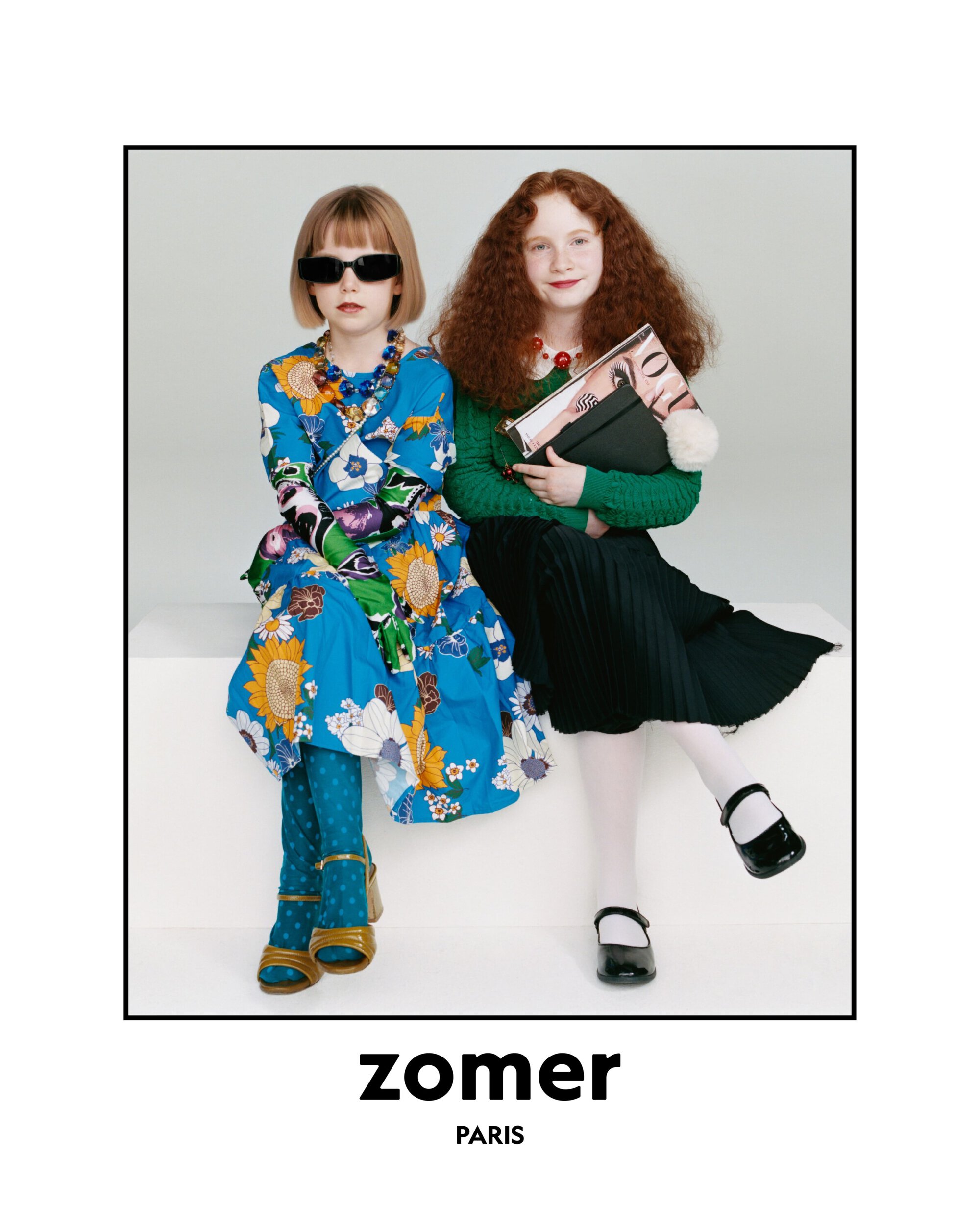 Two young girls in fashionable outfits sit in a photo studio. One girl wears a floral dress, the other a green sweater and black skirt.