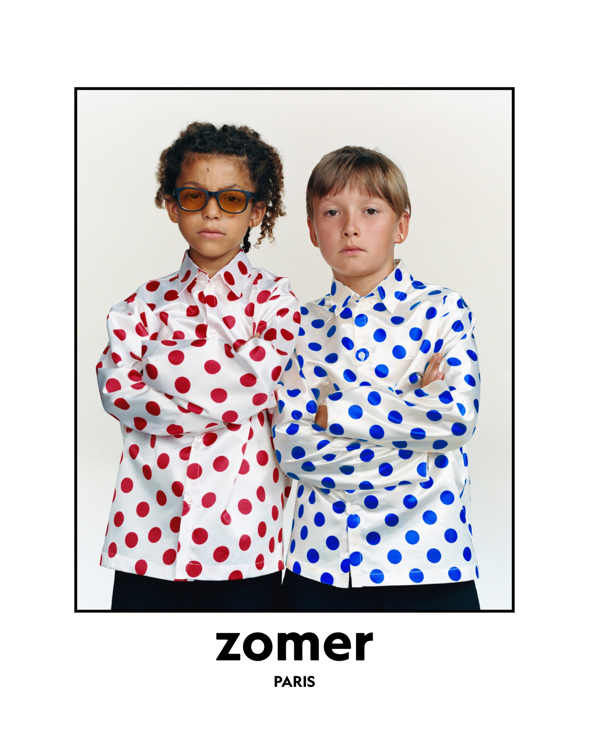 Two kids with serious expressions pose in white shirts with red and blue polka dots against a clean, bright studio backdrop.