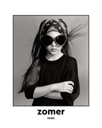 Black and white studio portrait of a young girl wearing oversized dark sunglasses, a paisley bandana, and a black sweater with crossed arms.