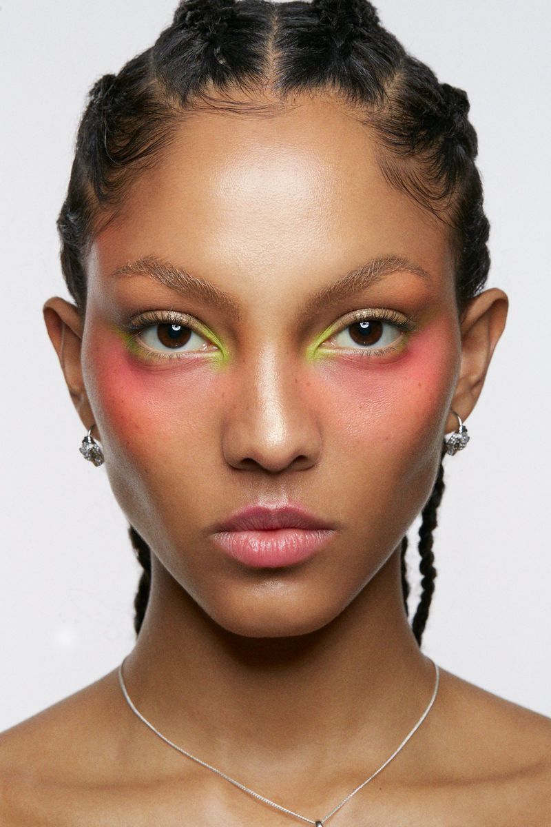 Close-up portrait of an adult woman with braided hair, wearing colorful makeup and silver jewelry against a plain white backdrop.