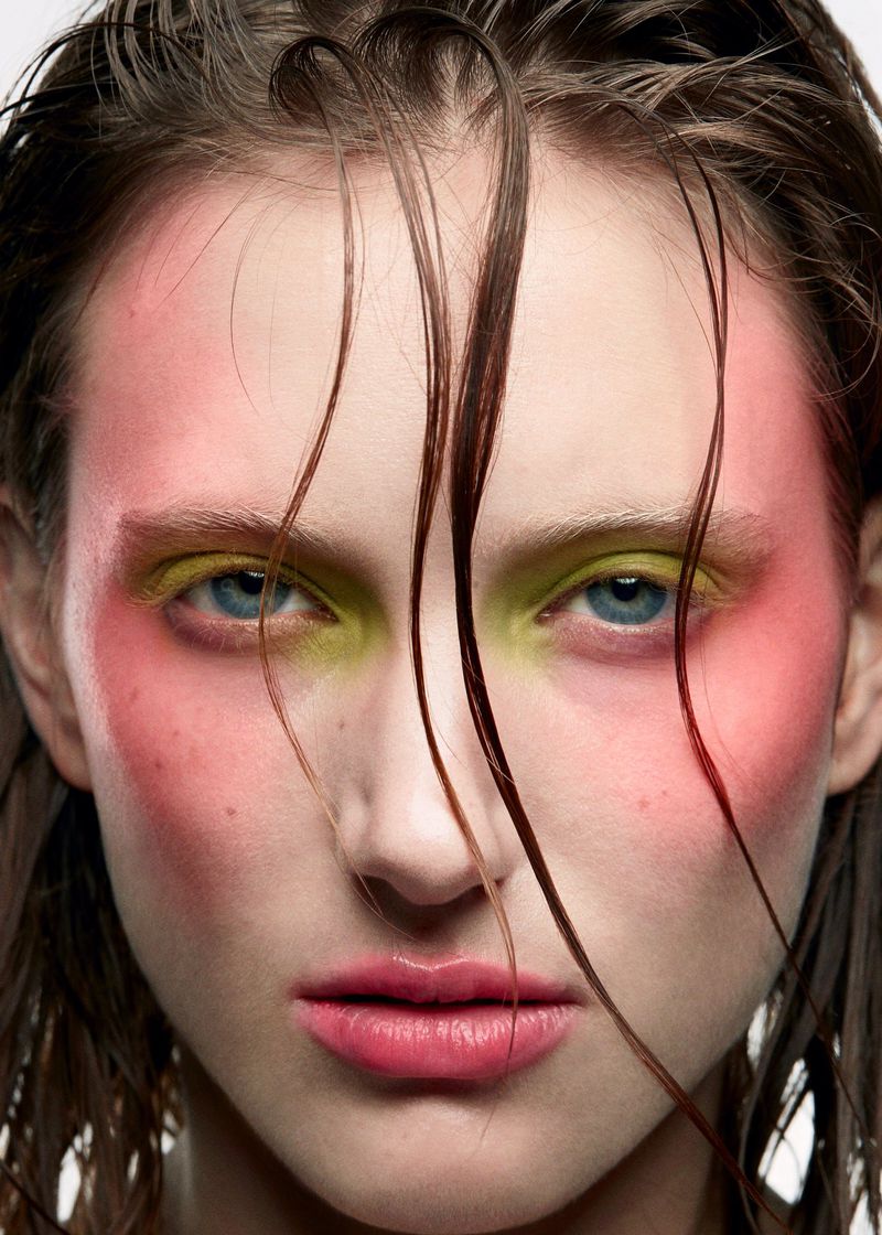 Close-up portrait of a woman with vibrant makeup featuring pink blush and bright yellow eyeshadow, highlighting the eyes and facial structure.