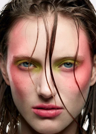 Close-up portrait of a woman with vibrant makeup featuring pink blush and bright yellow eyeshadow, highlighting the eyes and facial structure.