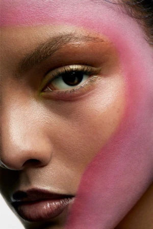 Close-up portrait of a woman with striking makeup featuring pink brushstrokes on her face, highlighting her eye and features against white.
