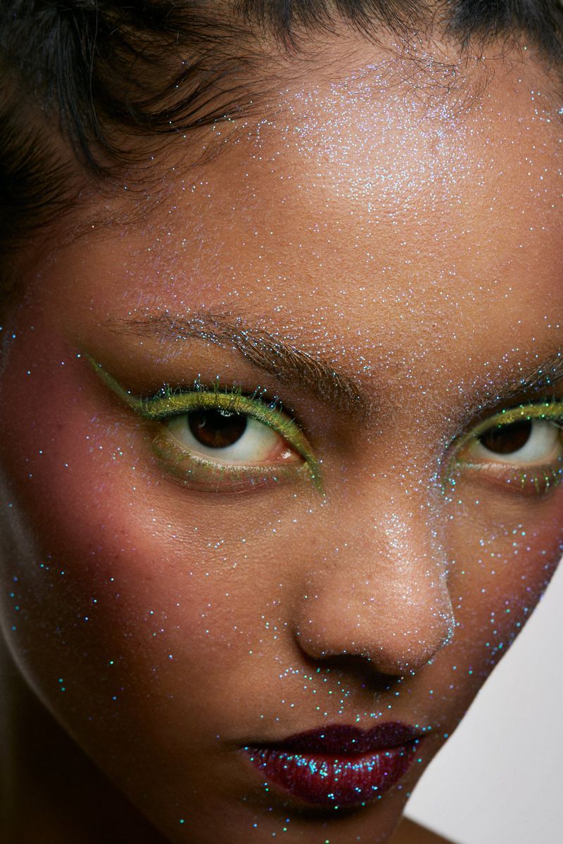 Closeup portrait of a woman's face with glitter makeup, including green eyeliner and iridescent glitter on her skin, set against a white backdrop.