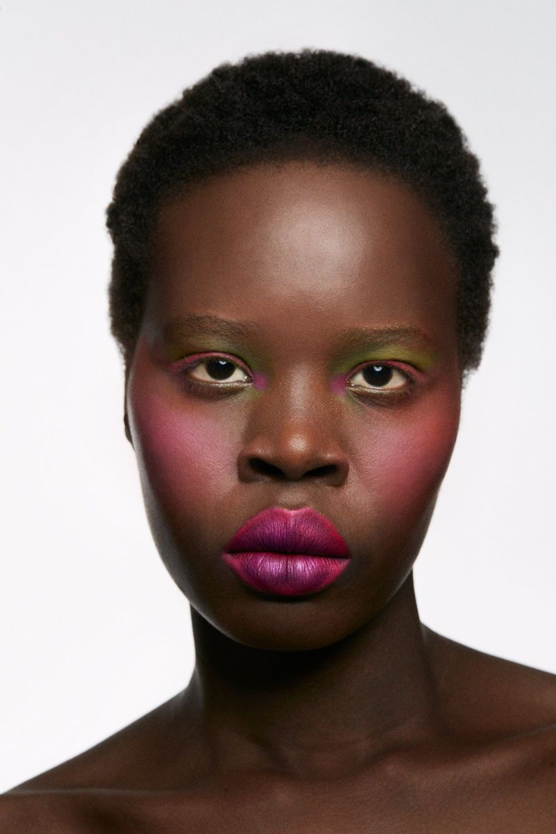 Close-up studio shot of a dark-skinned woman with vivid makeup: purple lips, pink cheeks, and green eyeshadow, set against a white background.
