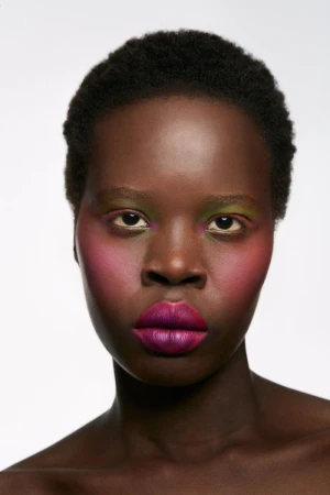 Close-up studio shot of a dark-skinned woman with vivid makeup: purple lips, pink cheeks, and green eyeshadow, set against a white background.