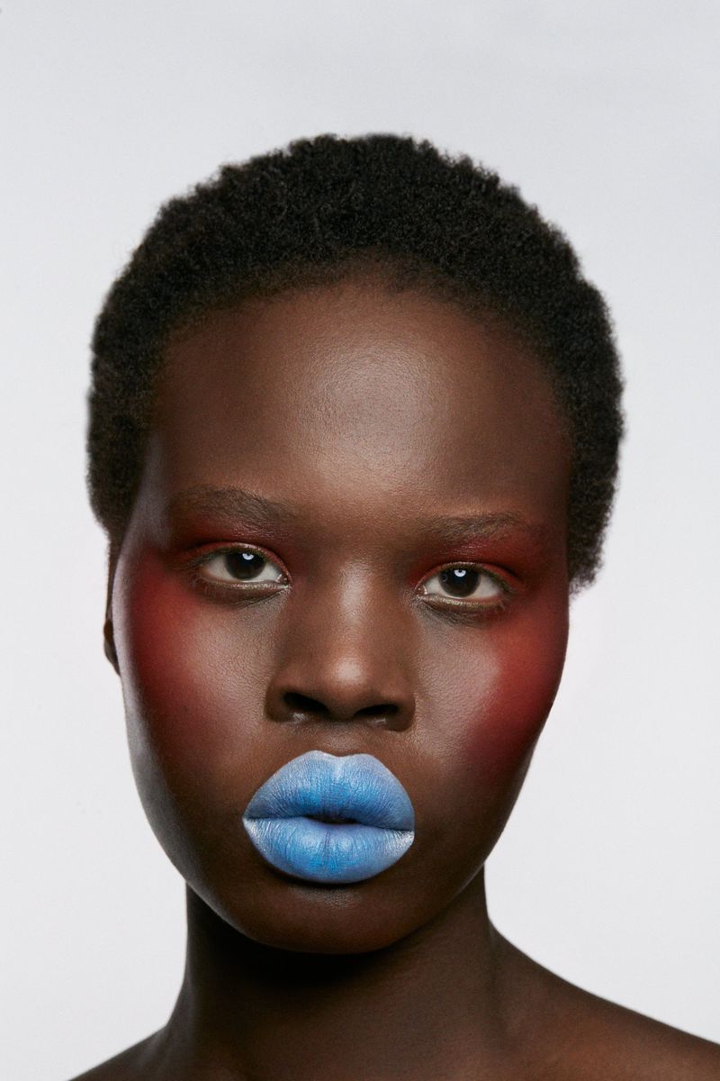 Close-up portrait of a woman with red makeup on her cheeks and blue lipstick against a white background in a beauty shot.