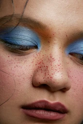 Close-up portrait of a model with vibrant blue eyeshadow, speckled red freckles on the nose, and dark mauve lips.