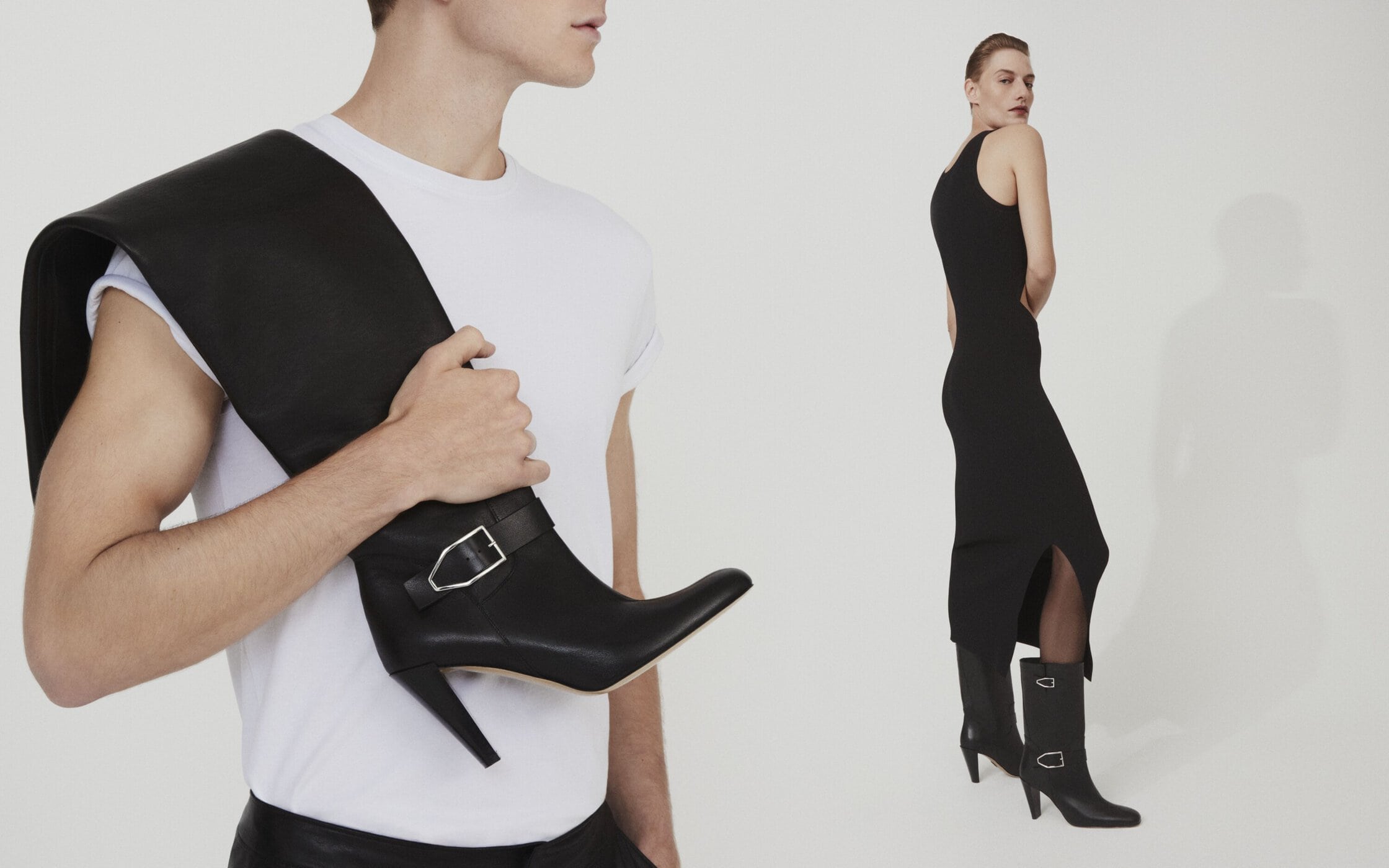 Two people in a studio setting, one carrying a high-heeled boot; the other modeling a black dress and matching heeled boots.