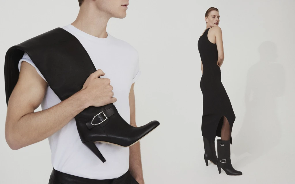 Two people in a studio setting, one carrying a high-heeled boot; the other modeling a black dress and matching heeled boots.