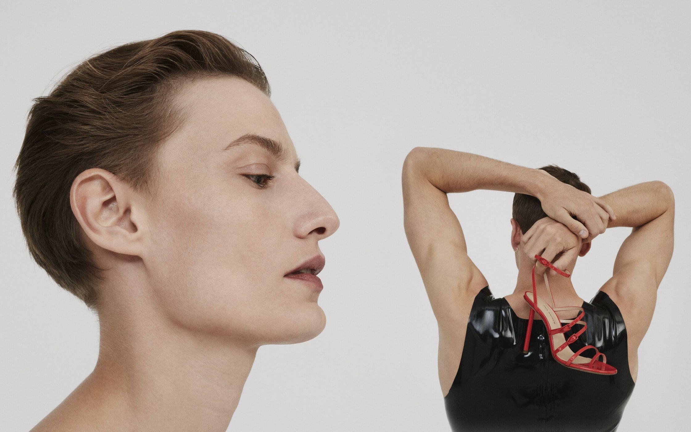 Close-up of two adult humans with contrasting presentation, one in makeup facing forward, the other holding red high-heel shoes, back to camera.