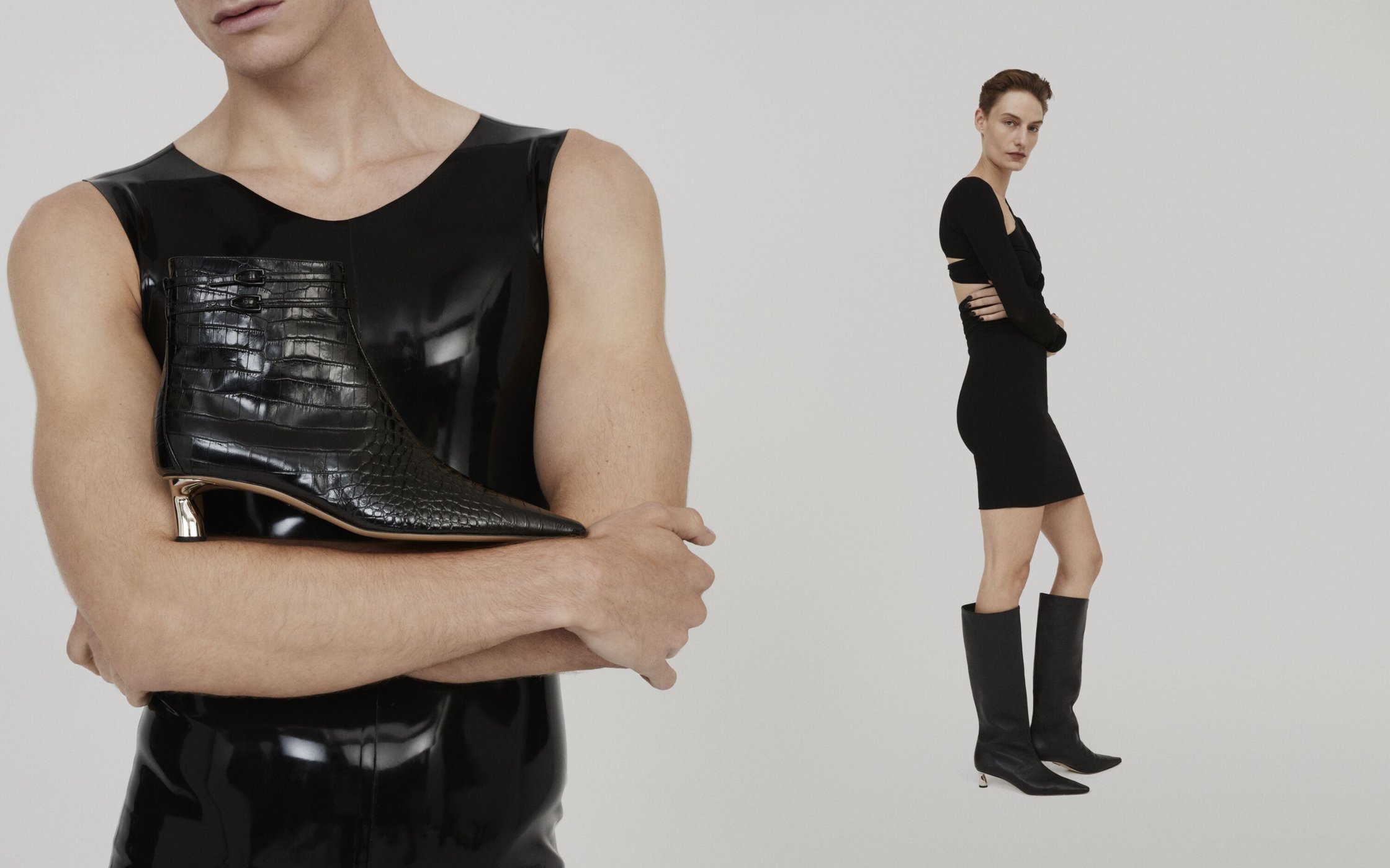 Fashion editorial shot of a man in a black latex top holding a black crocodile-print boot, and a woman in a black dress and boots.