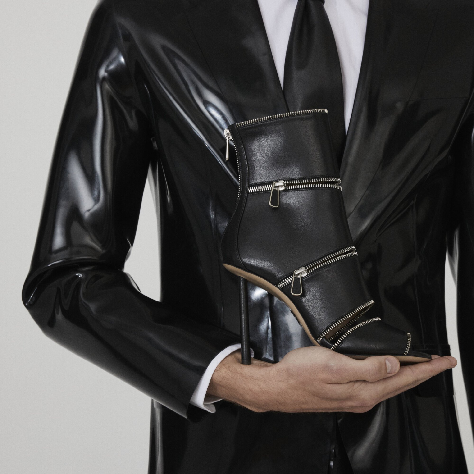 Black high-heeled boot with zipper detail held by a person wearing a shiny black jacket, white shirt, and tie against a grey background.