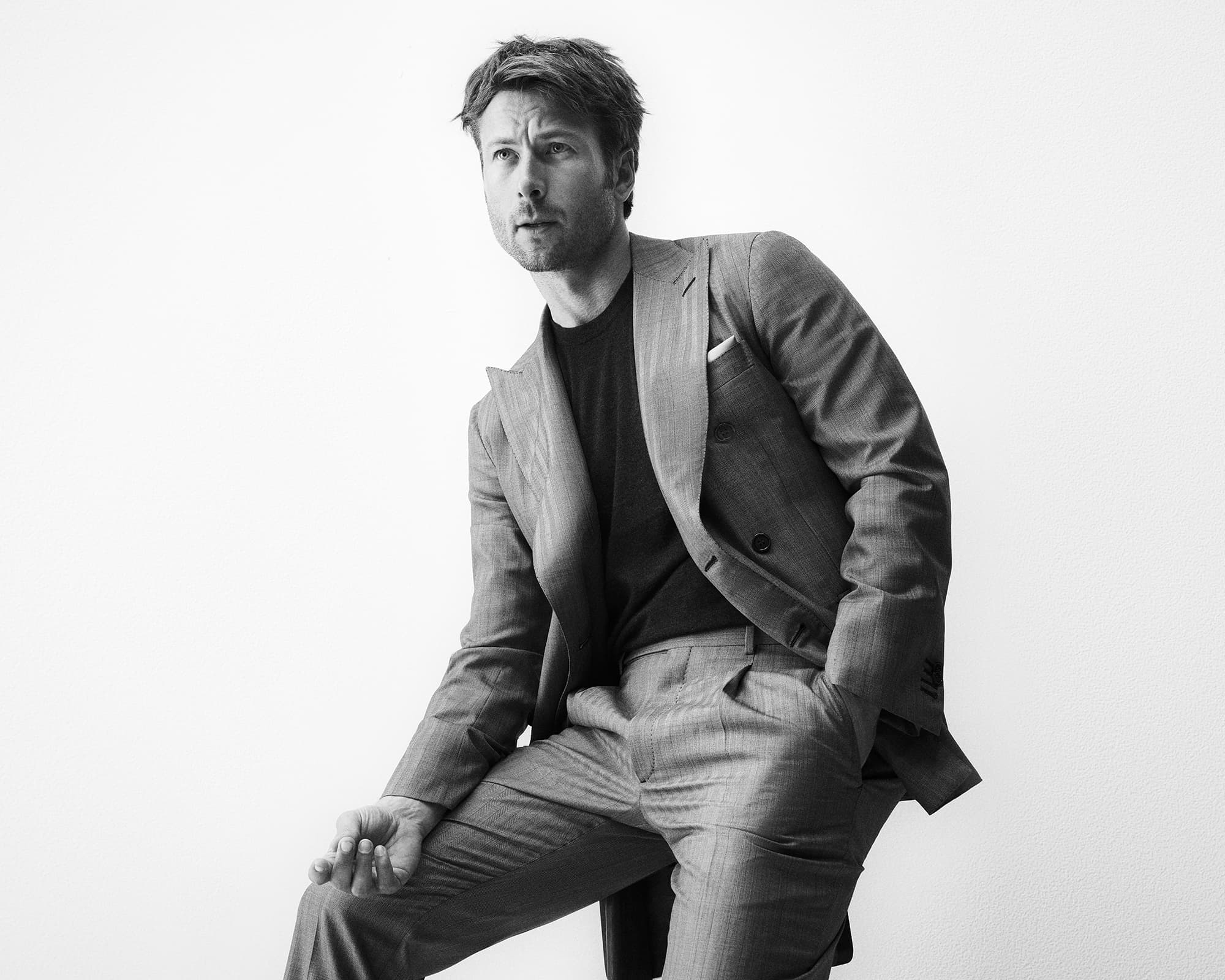 Man in a grey suit posing indoors against a white background, looking to the side with one hand in his pocket, shot in black and white.