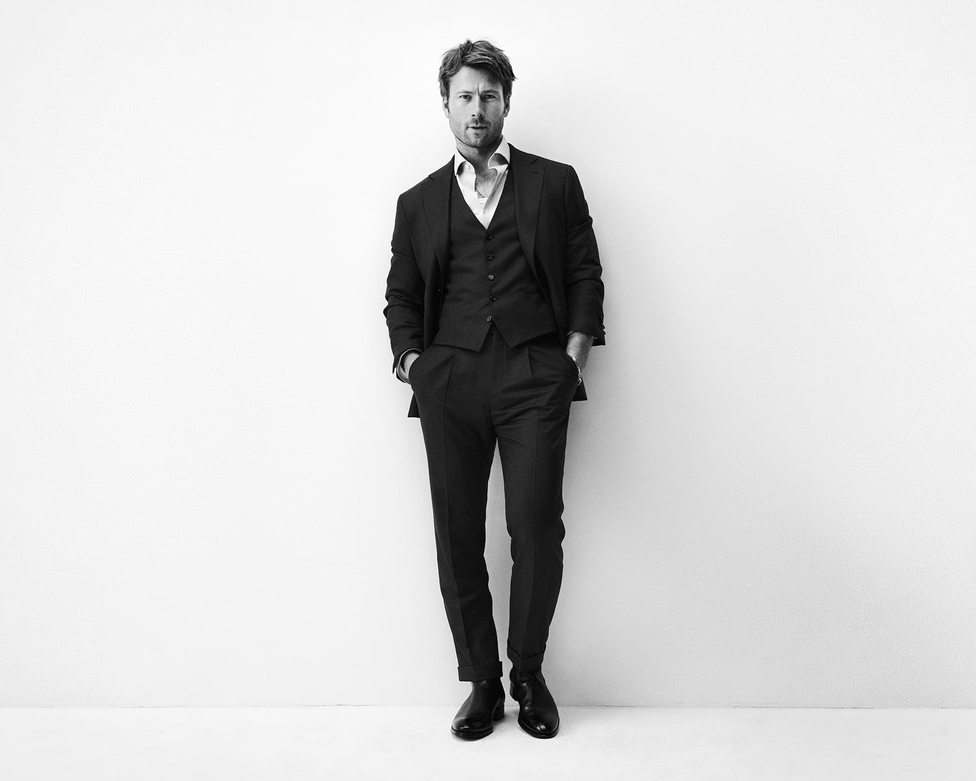 Full-length shot of a confident adult man in a dark suit, vest, and collared shirt, hands in pockets, leaning against a white wall.