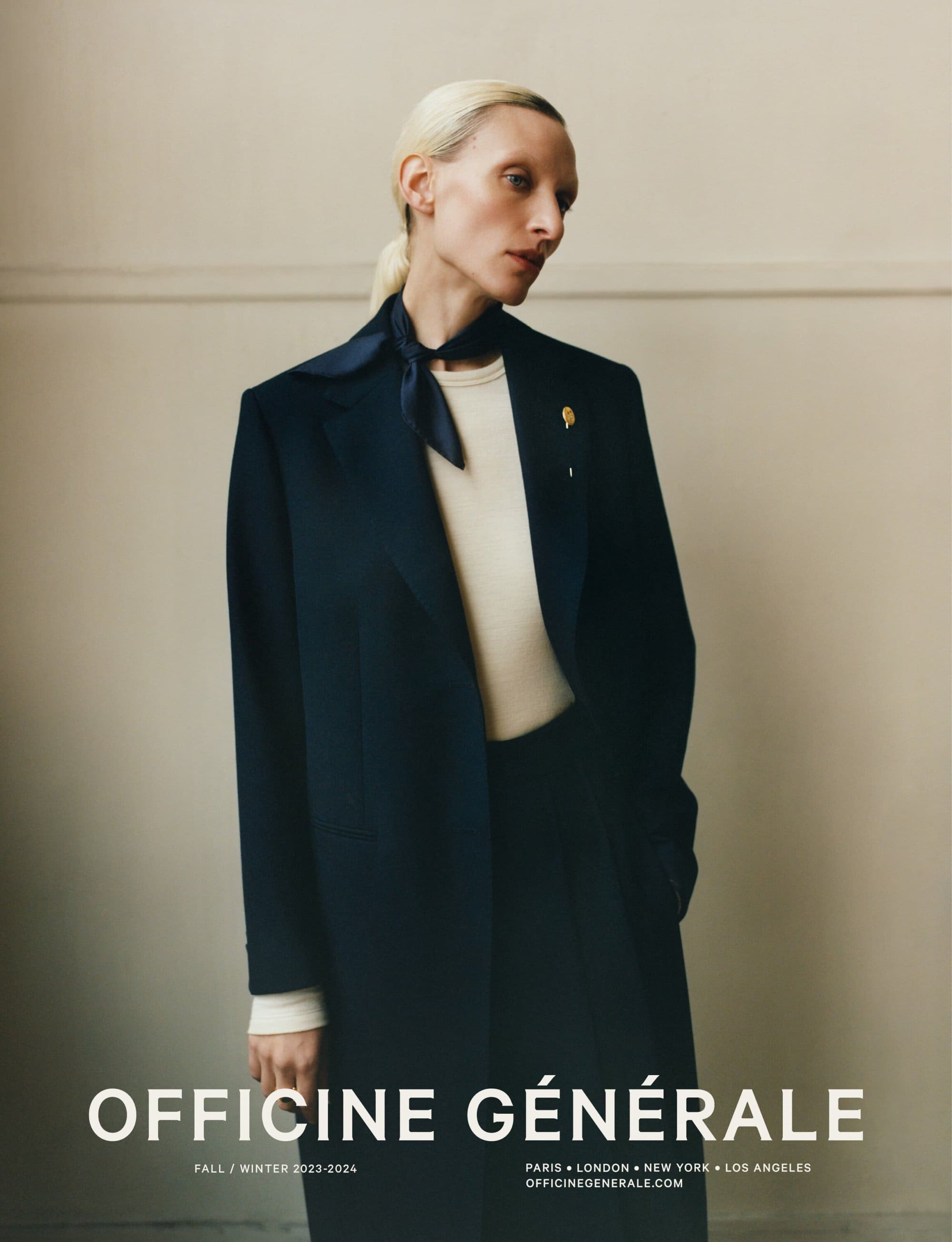 Model with blonde hair wearing a dark coat and neck scarf against a neutral wall for Officine Generale Fall 2023 campaign.
