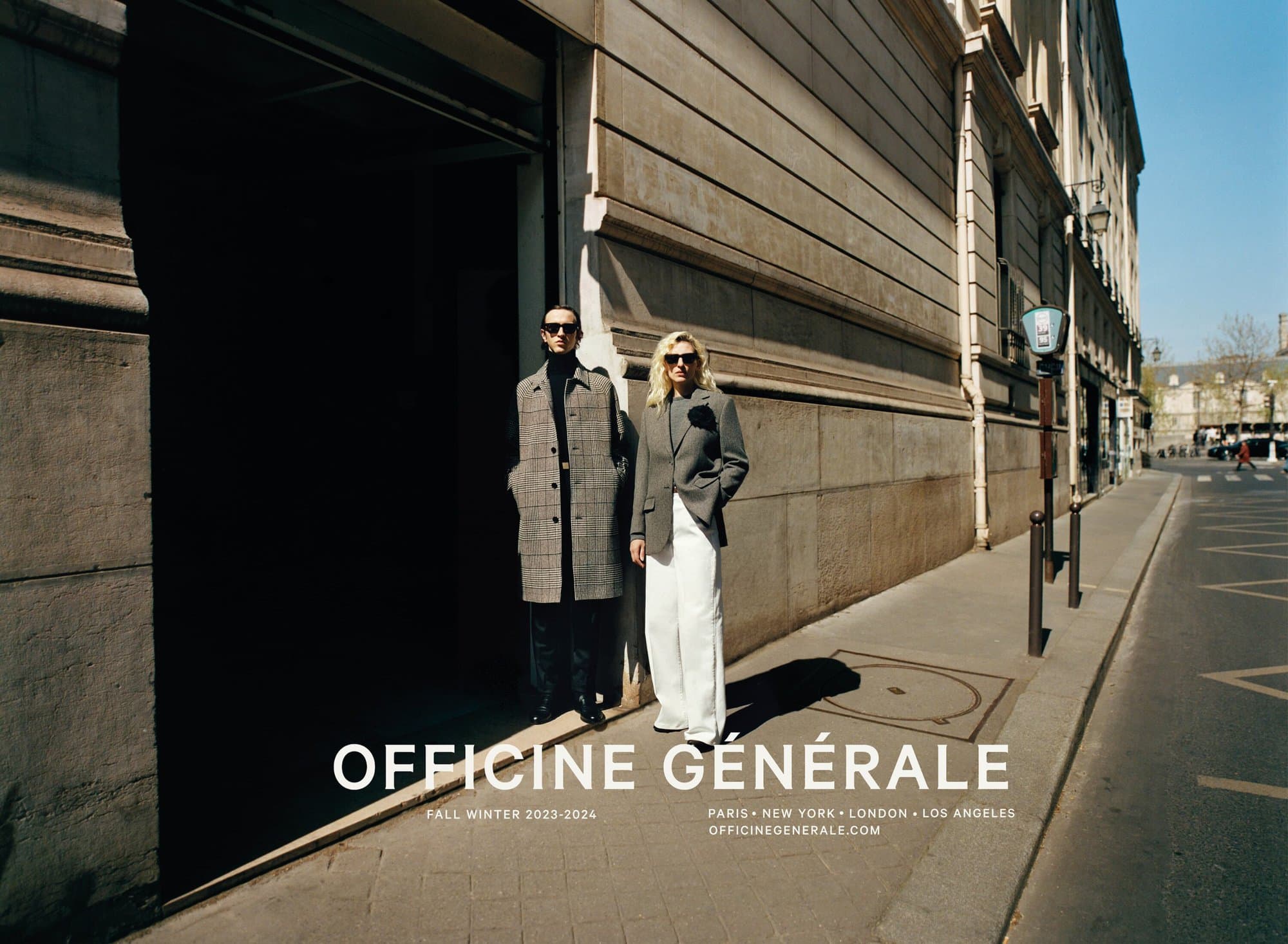 Two fashion models pose in gray jackets and pants on a sunny street in Paris, showcasing Fall Winter 2023-2024 collection.