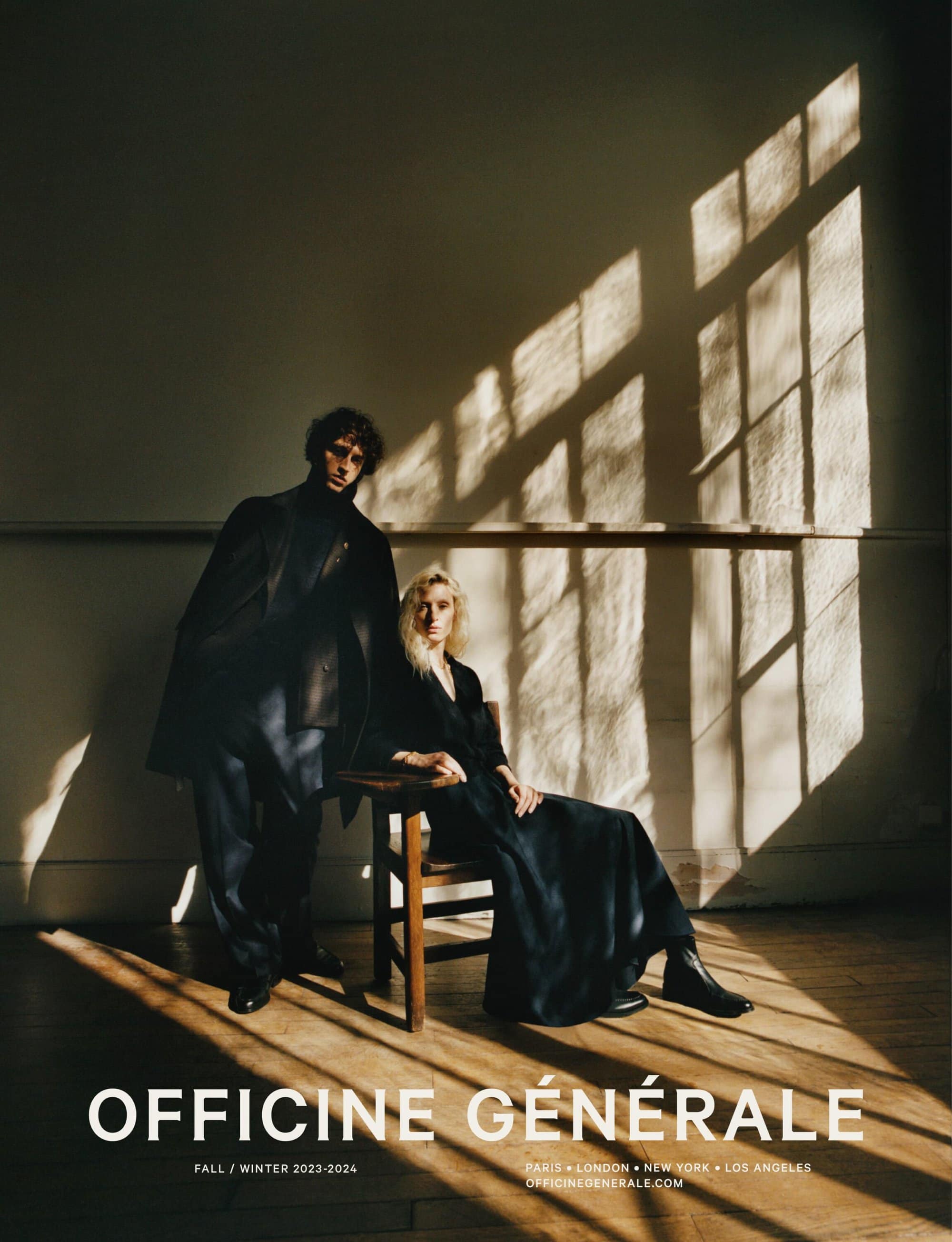 Fashion editorial shot of a man and woman indoors near a window with light patterns wearing dark clothing. 