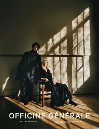 Fashion editorial shot of a man and woman indoors near a window with light patterns wearing dark clothing. 