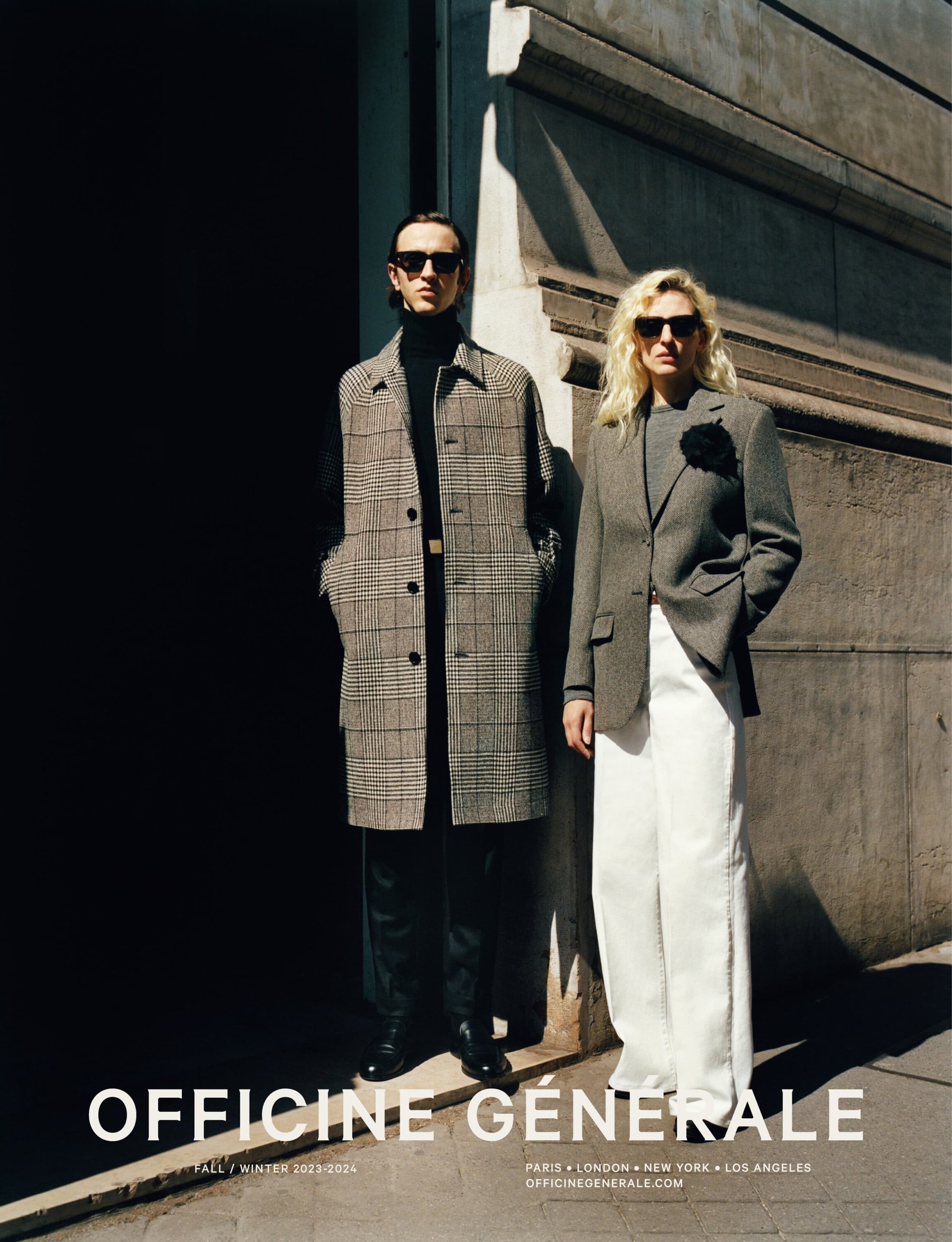 Fashionable adults in sunglasses pose outdoors near a building in an editorial style. One wears a plaid coat, the other a blazer and white pants.