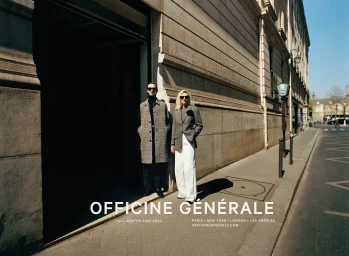 Two models standing on a city sidewalk in front of a stone building exterior wearing tailored coats and sunglasses.