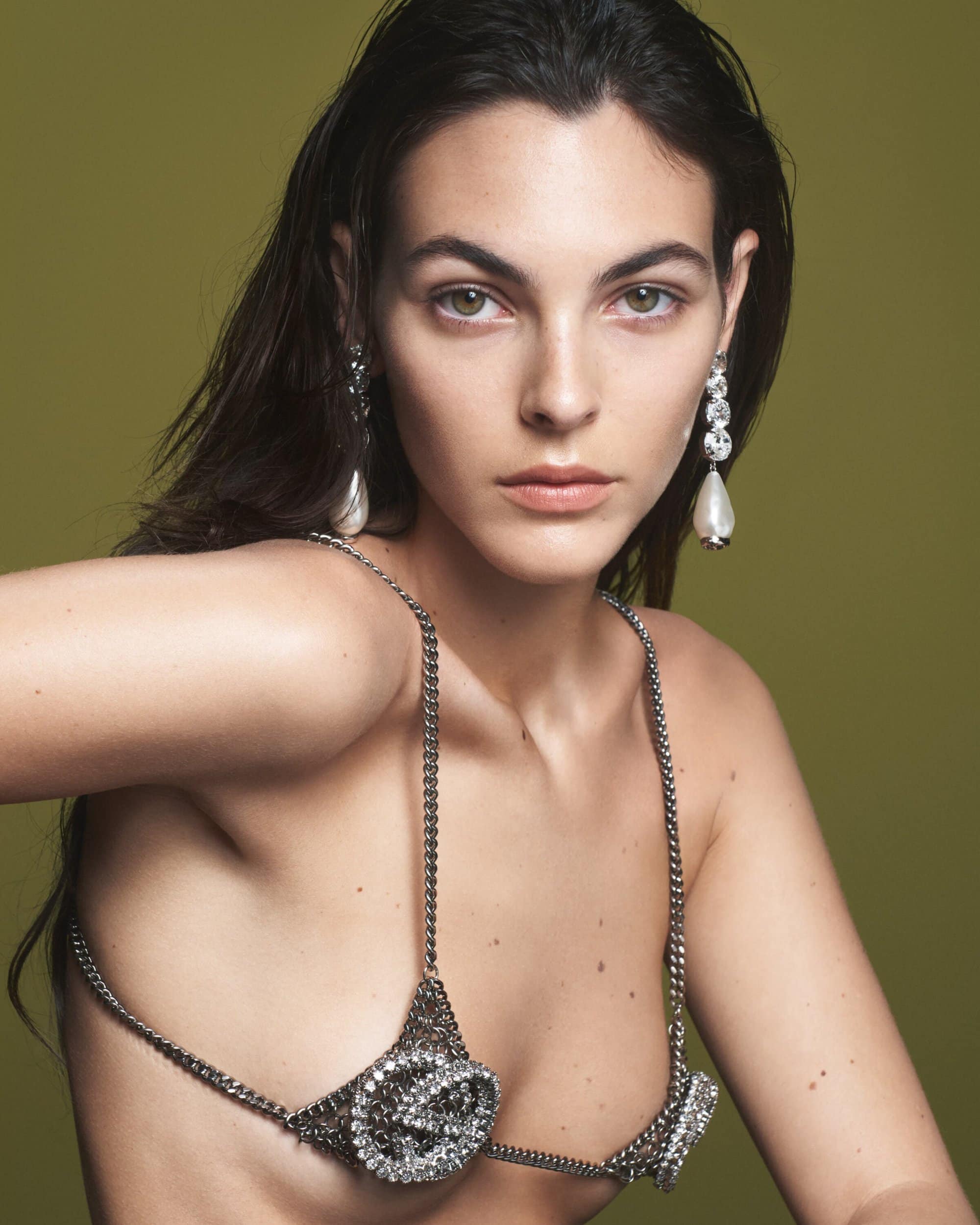 Brunette woman wearing a silver crystal chain bra and drop earrings poses in front of an olive green backdrop in a studio shot.