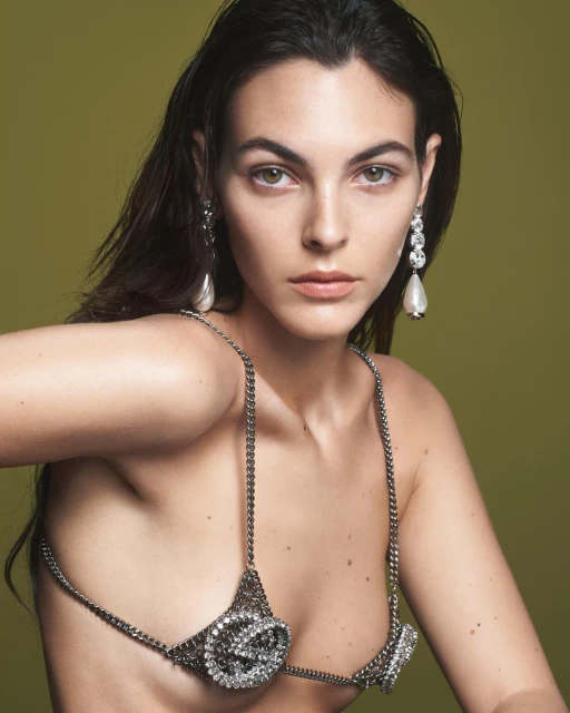 Brunette woman wearing a silver crystal chain bra and drop earrings poses in front of an olive green backdrop in a studio shot.