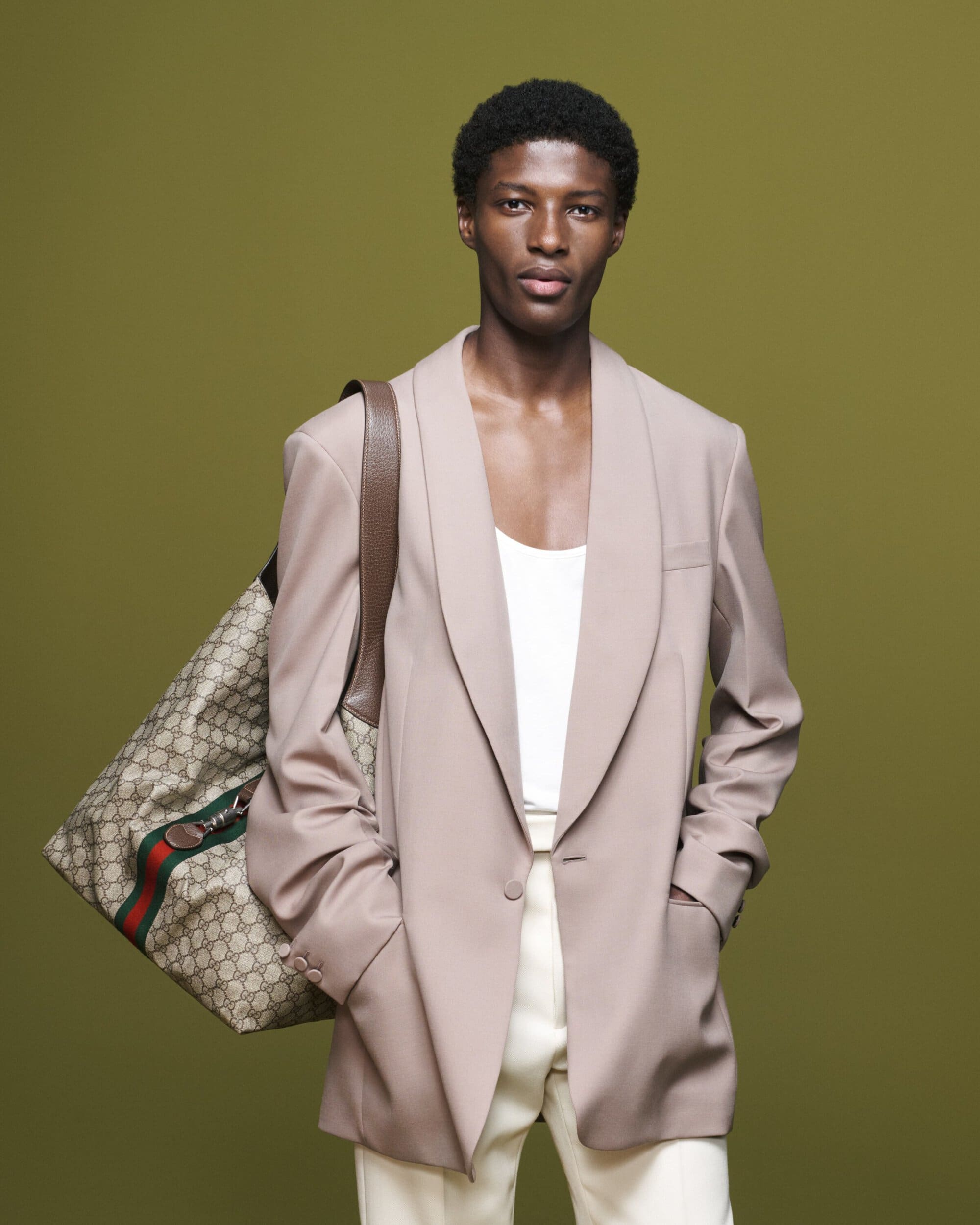 Fashionable African-American man in a blazer, white top, and light pants poses with a patterned Gucci bag against a green backdrop.