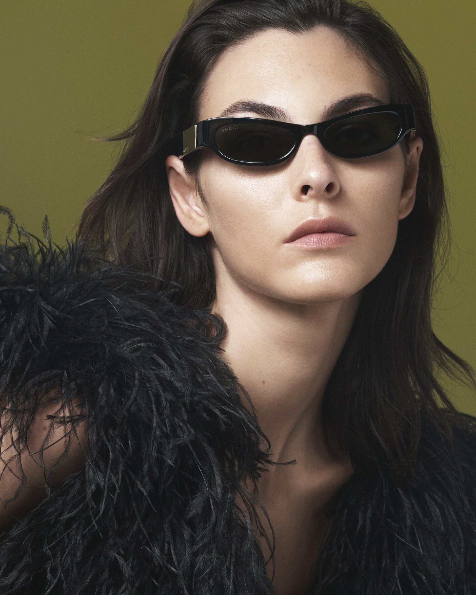 Close-up of a woman wearing sunglasses and a black feathered garment against an olive-green backdrop in a fashion-style portrait.