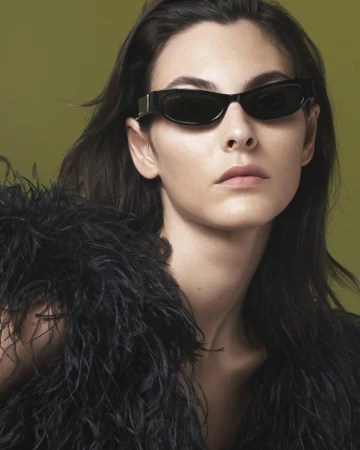 Close-up of a woman wearing sunglasses and a black feathered garment against an olive-green backdrop in a fashion-style portrait.