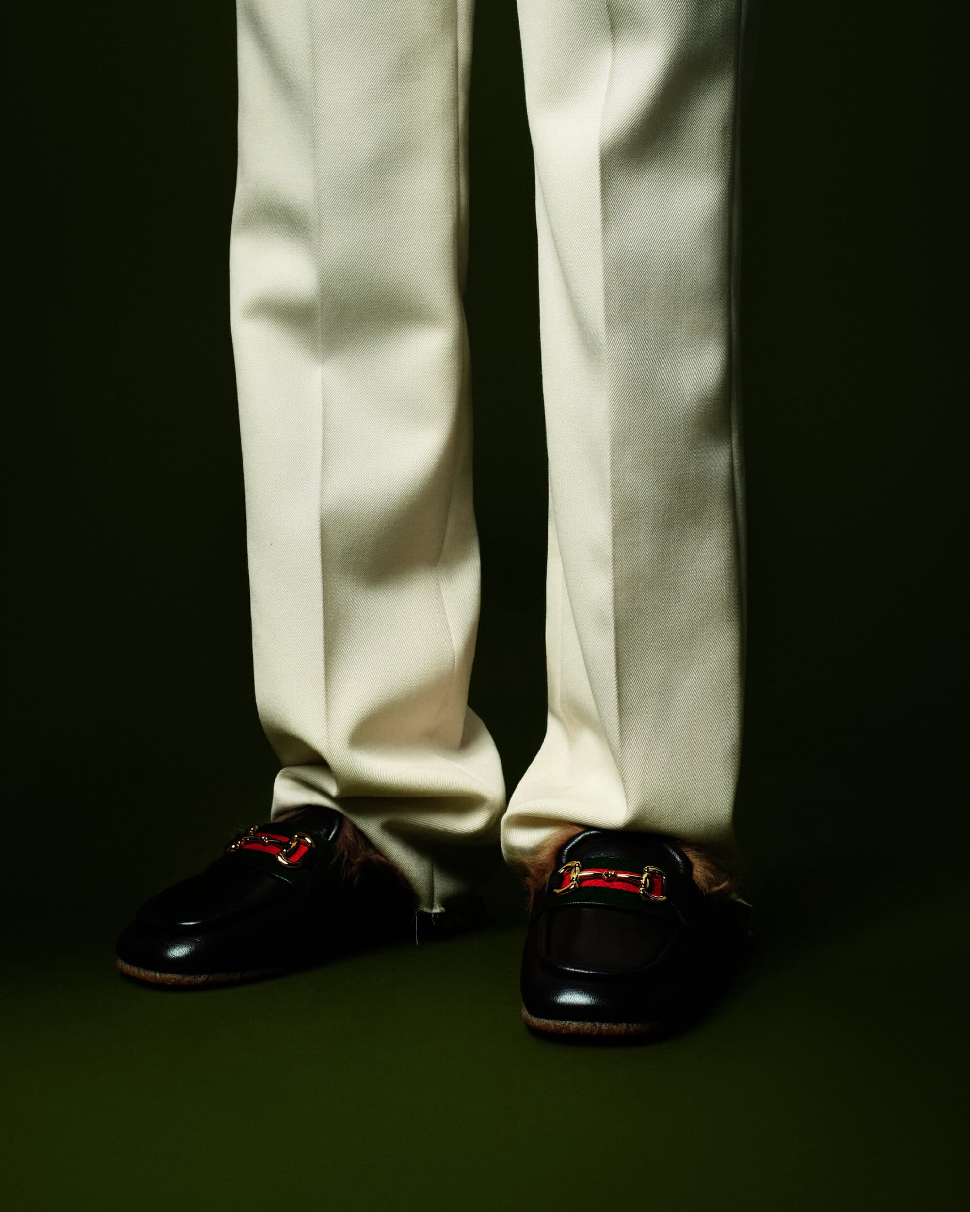Close-up of a person wearing cream-colored trousers and black loafers with gold hardware, set against a dark green background.