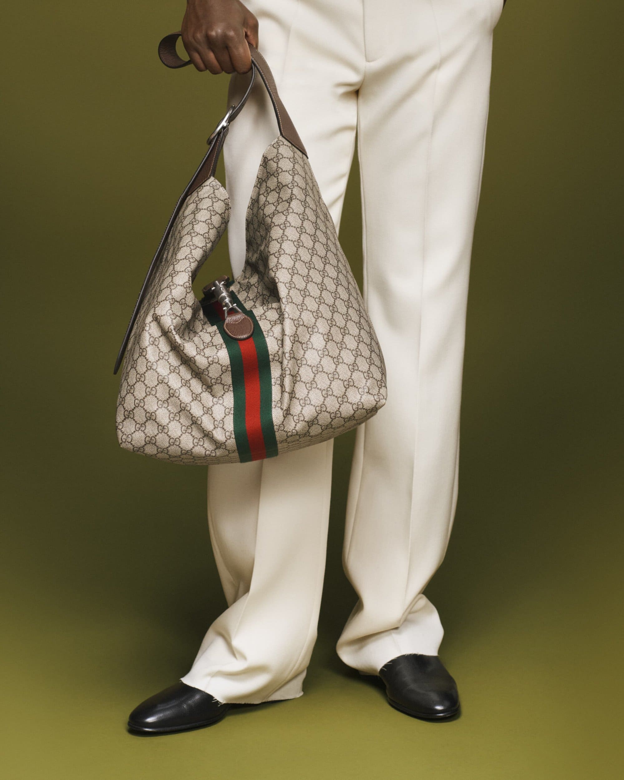 Person wearing white trousers holding a monogram canvas shoulder bag with a red and green stripe