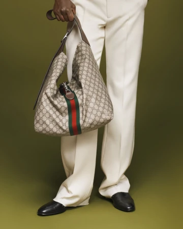 Person wearing white trousers holding a monogram canvas shoulder bag with a red and green stripe