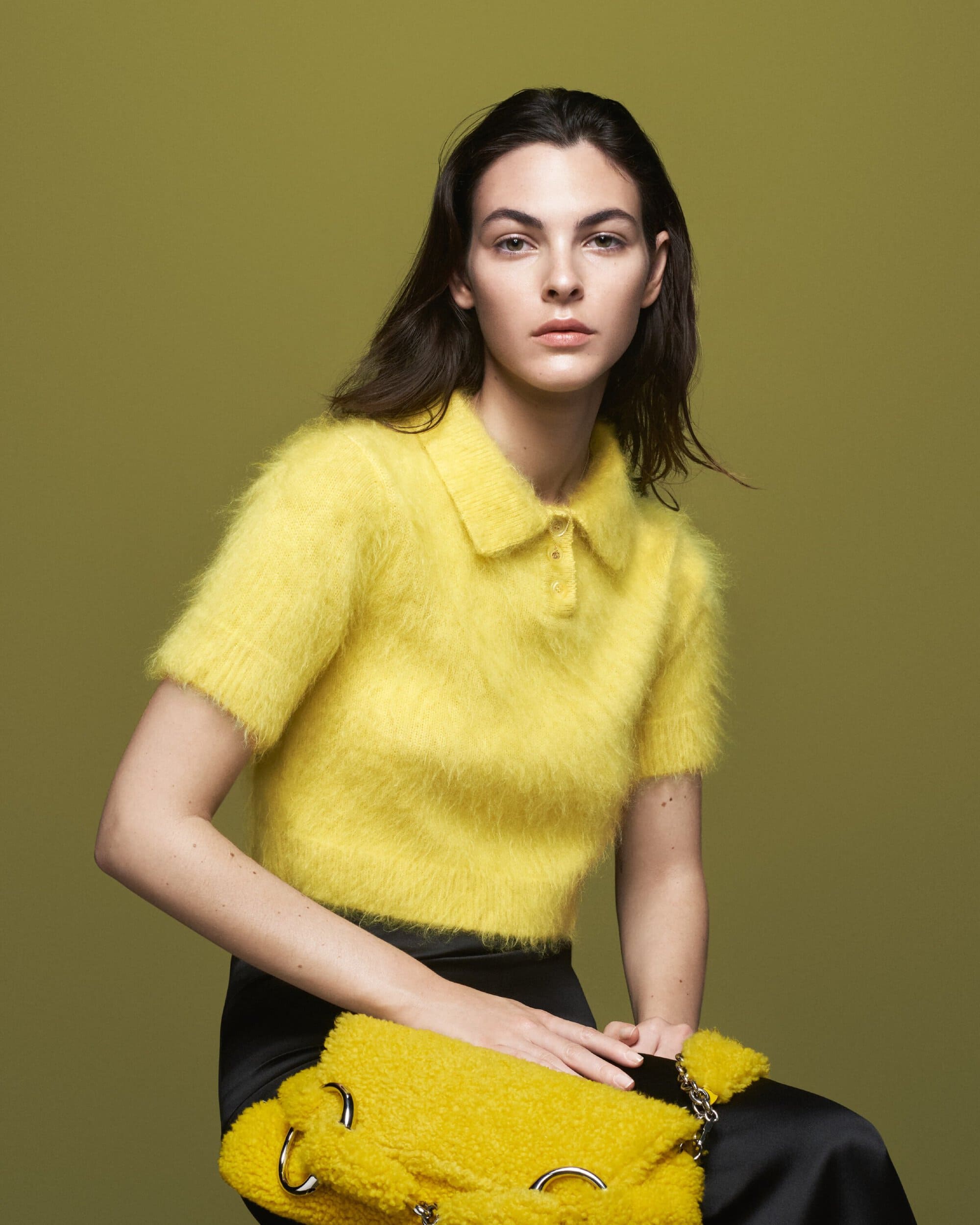 A brunette woman poses in a studio with a yellow fuzzy collared shirt, black skirt, and matching yellow bag against an olive backdrop.
