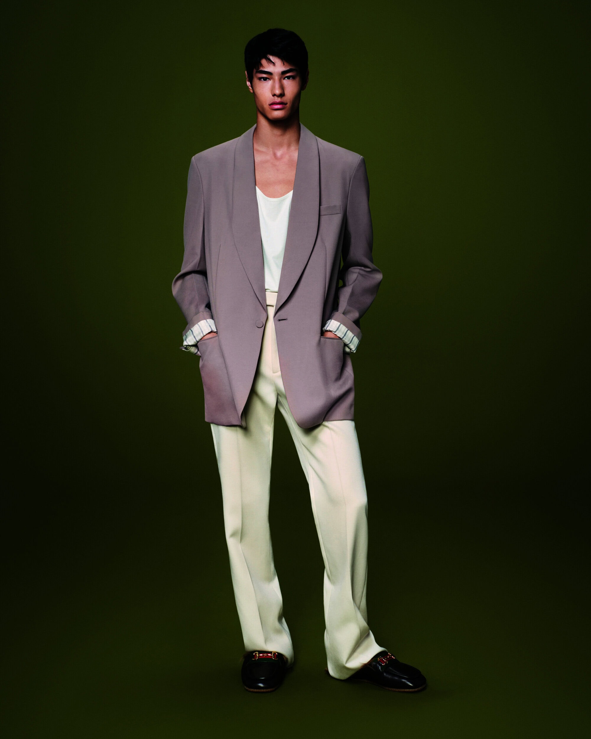 Young man in a taupe blazer with white interior lining, white tank top, and light trousers standing against a deep forest green background.