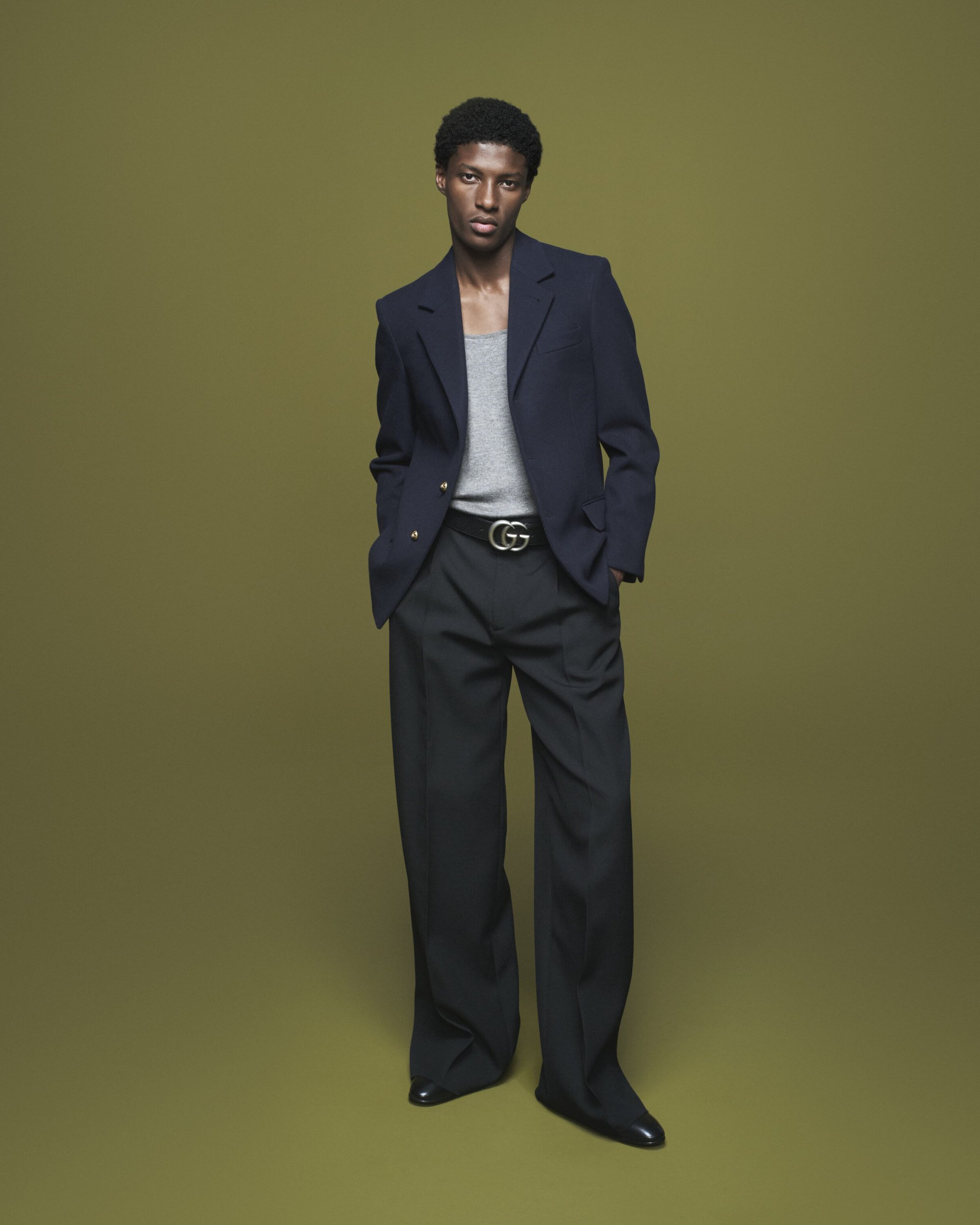 Full-length portrait of a stylish Black man in a navy blazer, gray tank top, and black trousers against a muted olive green background.