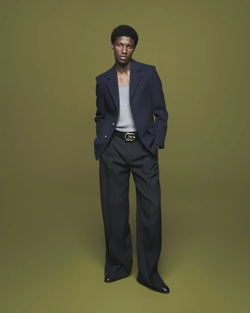 Full-length portrait of a stylish Black man in a navy blazer, gray tank top, and black trousers against a muted olive green background.