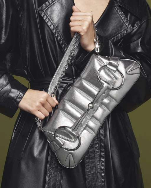 A shiny silver shoulder bag with a metal horse-bit detail is held by a woman in a black leather trench coat against a muted green backdrop.