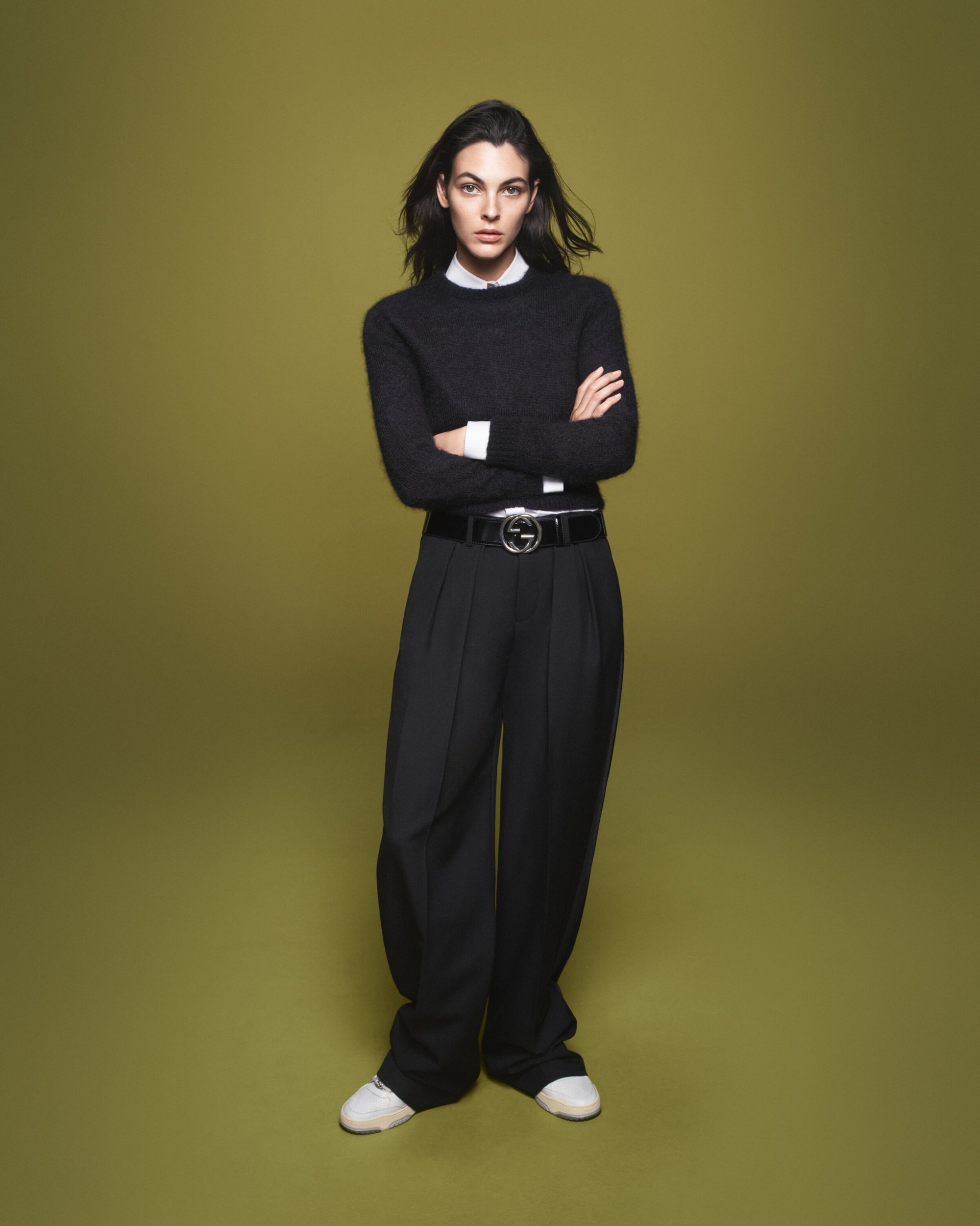 Full-length portrait of a woman with dark hair wearing black sweater, wide-legged black pants, Gucci belt and white sneakers against olive-green background.