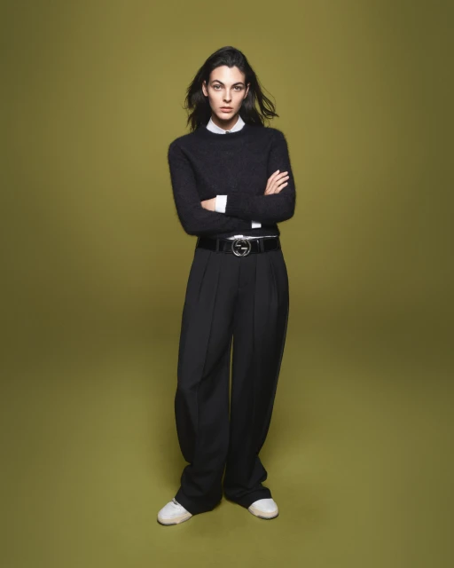 Full-length portrait of a woman with dark hair wearing black sweater, wide-legged black pants, Gucci belt and white sneakers against olive-green background.