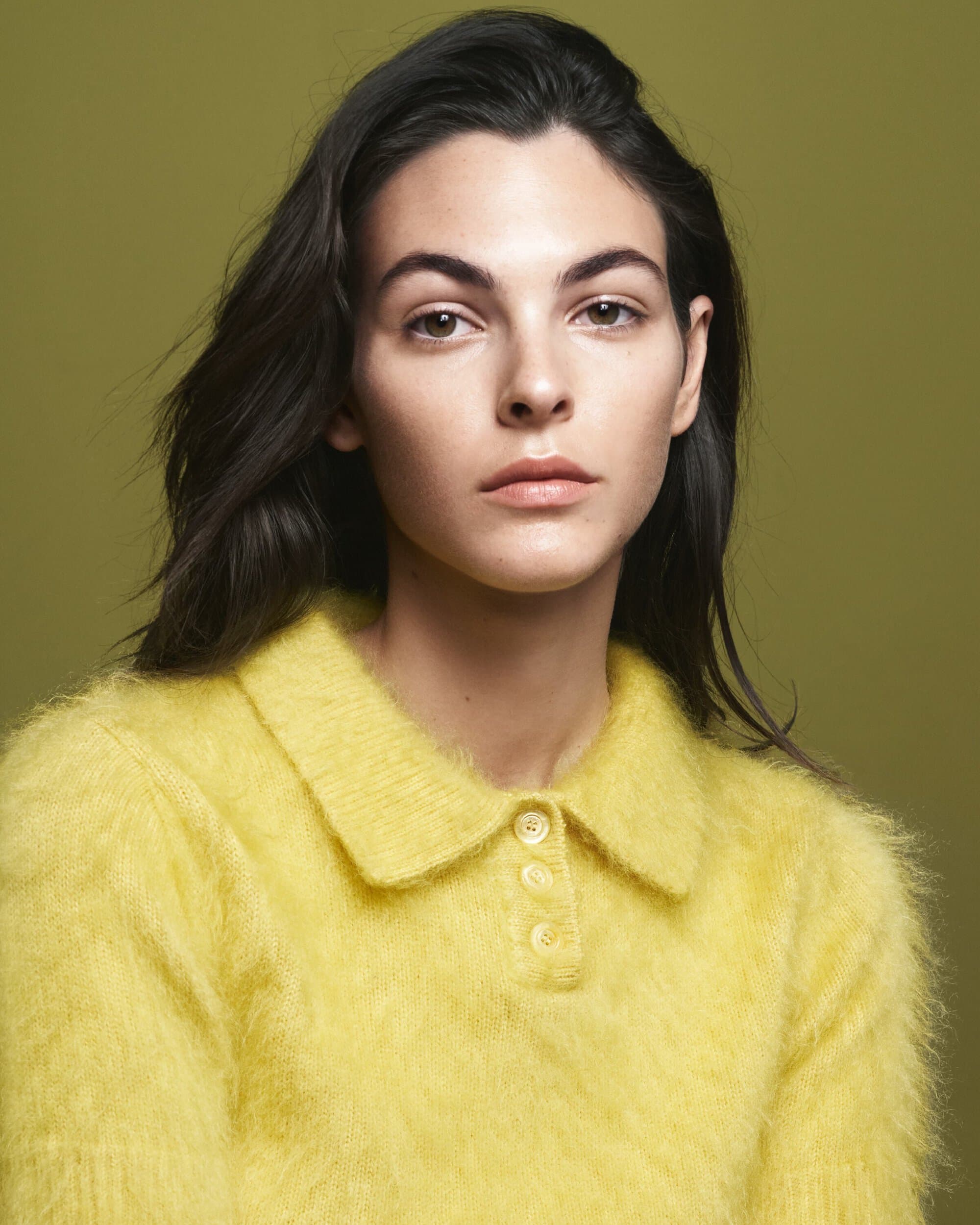 Close-up portrait of a young brunette woman with fair skin wearing a yellow sweater against a soft olive green background in studio.